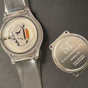 May include: A clear plastic wristwatch with the back cover removed, revealing the internal components. The watch face is white, and the watch band is transparent. The detached back cover is silver and has text that reads "The Metropolitan Museum of Art, MMA.CN, Water Resistant, Japan Movement."