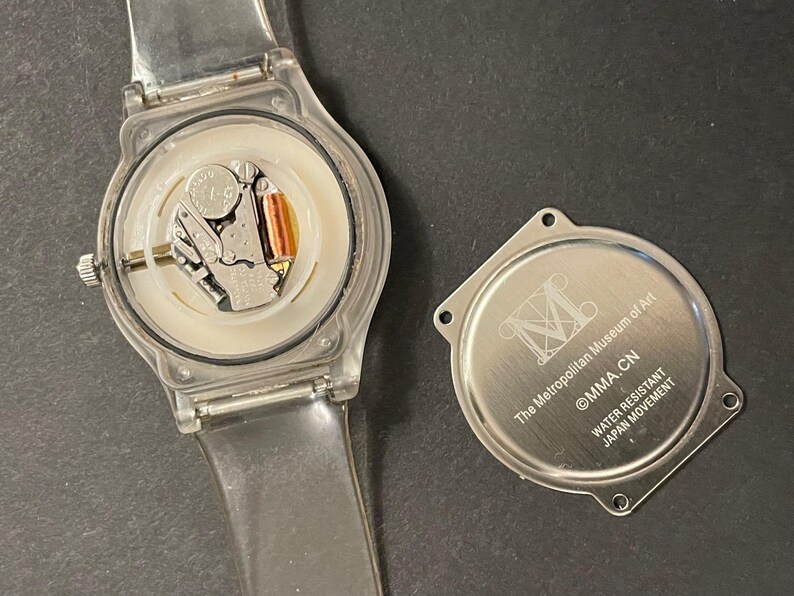 May include: A clear plastic wristwatch with the back cover removed, revealing the internal components. The watch face is white, and the watch band is transparent. The detached back cover is silver and has text that reads "The Metropolitan Museum of Art, MMA.CN, Water Resistant, Japan Movement."
