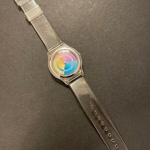 May include: Clear plastic wristwatch with a colorful circular face. The watch face features segments of yellow, orange, purple, and blue. The clear band has a buckle closure and is approximately 9.5 inches long.