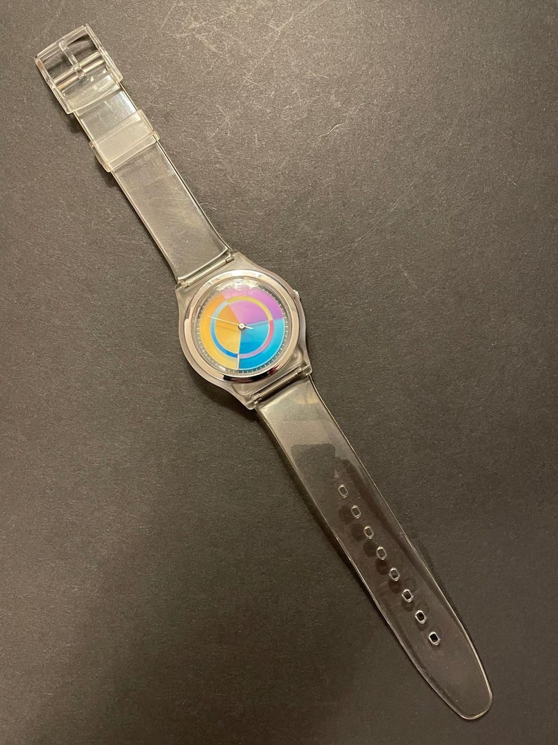 May include: Clear plastic wristwatch with a colorful circular face. The watch face features segments of yellow, orange, purple, and blue. The clear band has a buckle closure and is approximately 9.5 inches long.