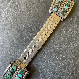 Op de afbeelding: Vintage zilverkleurige horlogeband met turquoise en groene stenen accenten. De rechthoekige uiteinden hebben twee rijen stenen, afwisselend turquoise en groen. De rekbare band heeft een rasterpatroon.