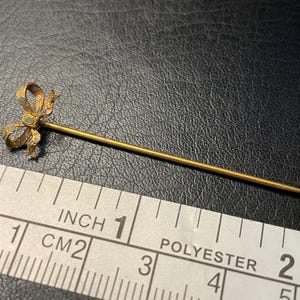 May include: A gold-coloured stick pin with a bow-shaped head. The bow has a detailed design with small, light-coloured accents. The pin is approximately 2.5 cm long.