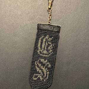 May include: A black beaded fob with gold-colored lettering and a gold-tone clasp. The fob is rectangular with a rounded bottom and a black ribbon at the top. The lettering appears to spell out a word or initials.