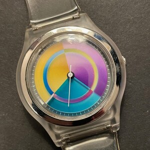 May include: A clear plastic wristwatch with a colorful dial. The dial features a segmented design with yellow, teal, purple, and pink sections. The watch has a silver-colored bezel and a clear plastic strap.