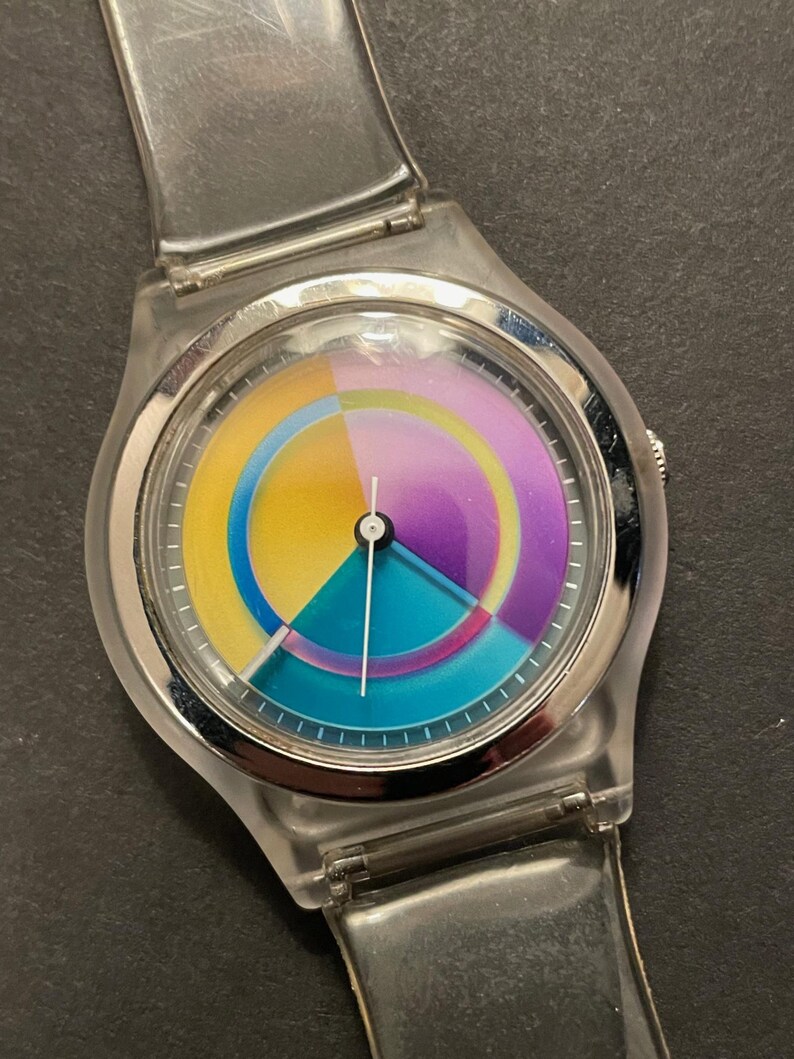 May include: A clear plastic wristwatch with a colorful dial. The dial features a segmented design with yellow, teal, purple, and pink sections. The watch has a silver-colored bezel and a clear plastic strap.