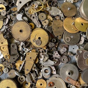 Könnte beinhalten: Nahaufnahme einer Sammlung von Vintage-Uhrenteilen, darunter Zahnräder, Platten und andere Komponenten. Die Teile bestehen aus Messing und Stahl, mit Gold- und Silbertönen. Die Anordnung ist dicht und detailliert.
