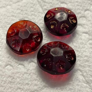May include: Three round, translucent, deep red glass buttons featuring a faceted top surface. Each button is decorated with a repeating embossed floral pattern highlighted in gold paint, arranged in a circular design around the centre.