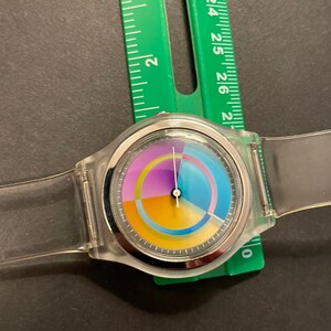 May include: A clear plastic wristwatch with a colorful segmented dial. The dial features sections of yellow, orange, purple, and blue. The watch is approximately 1.5 inches in diameter and has a clear plastic band. A green ruler is next to the watch.
