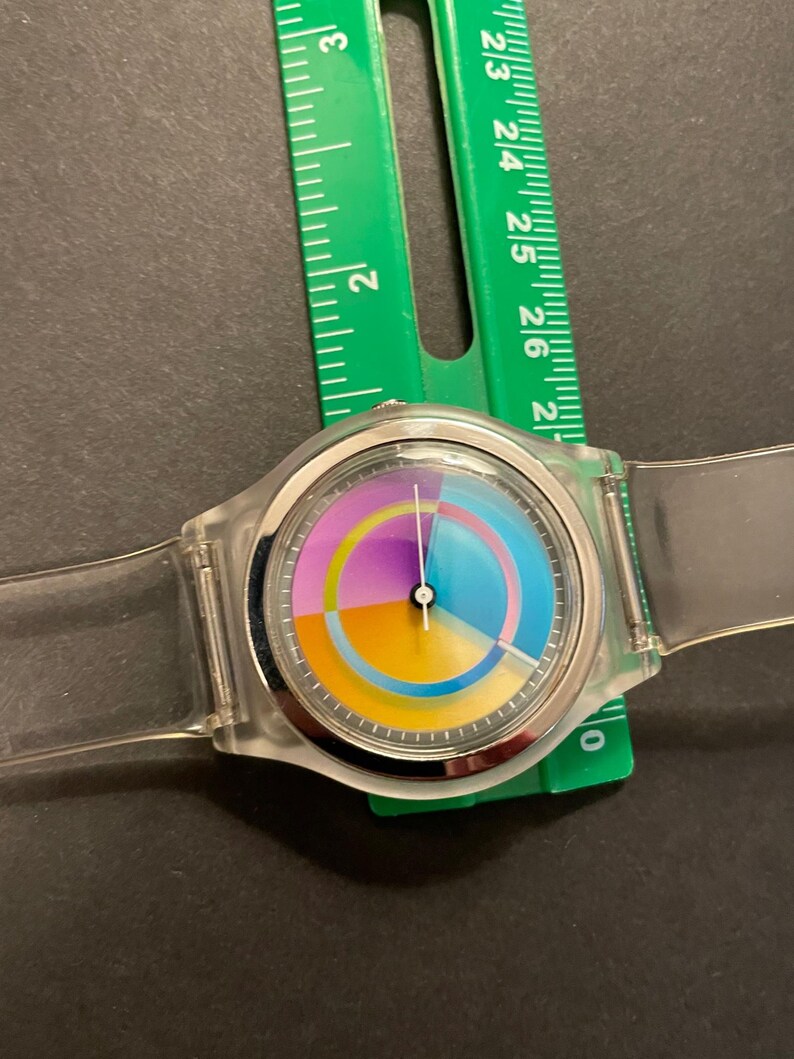 May include: A clear plastic wristwatch with a colorful segmented dial. The dial features sections of yellow, orange, purple, and blue. The watch is approximately 1.5 inches in diameter and has a clear plastic band. A green ruler is next to the watch.