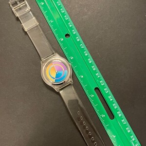 May include: A clear plastic wristwatch with a colorful rainbow-colored face and a clear plastic band. A green ruler is next to the watch. The ruler is marked with inches and has the word "CHINA" printed on it.