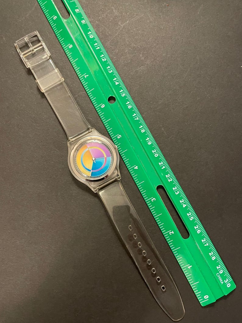 May include: A clear plastic wristwatch with a colorful rainbow-colored face and a clear plastic band. A green ruler is next to the watch. The ruler is marked with inches and has the word "CHINA" printed on it.