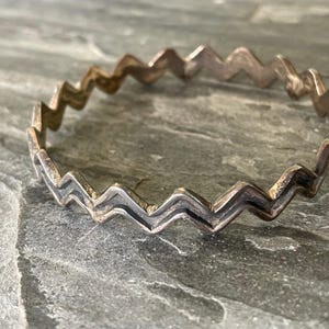 Puede incluir: Una pulsera rígida de metal en tono plateado que presenta un patrón continuo de zigzag o chevrón. La pulsera tiene un acabado pulido con áreas hundidas oscurecidas que resaltan los picos y valles angulares del diseño geométrico.