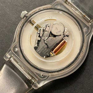 May include: A clear plastic watch with the back removed, revealing the internal mechanism. The watch face is white with a silver-colored metal movement and a small AG4 battery. The watch band is also clear plastic.