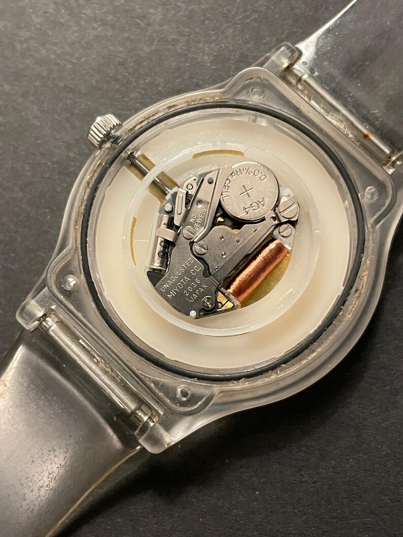 May include: A clear plastic watch with the back removed, revealing the internal mechanism. The watch face is white with a silver-colored metal movement and a small AG4 battery. The watch band is also clear plastic.