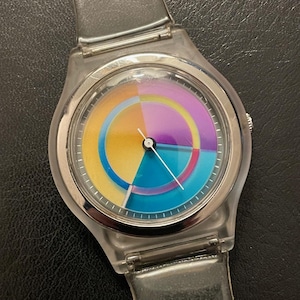 May include: A clear plastic wristwatch with a colorful, segmented dial. The dial features a vibrant array of colors including yellow, blue, purple, and orange. The watch has a silver-colored bezel and a clear plastic strap.