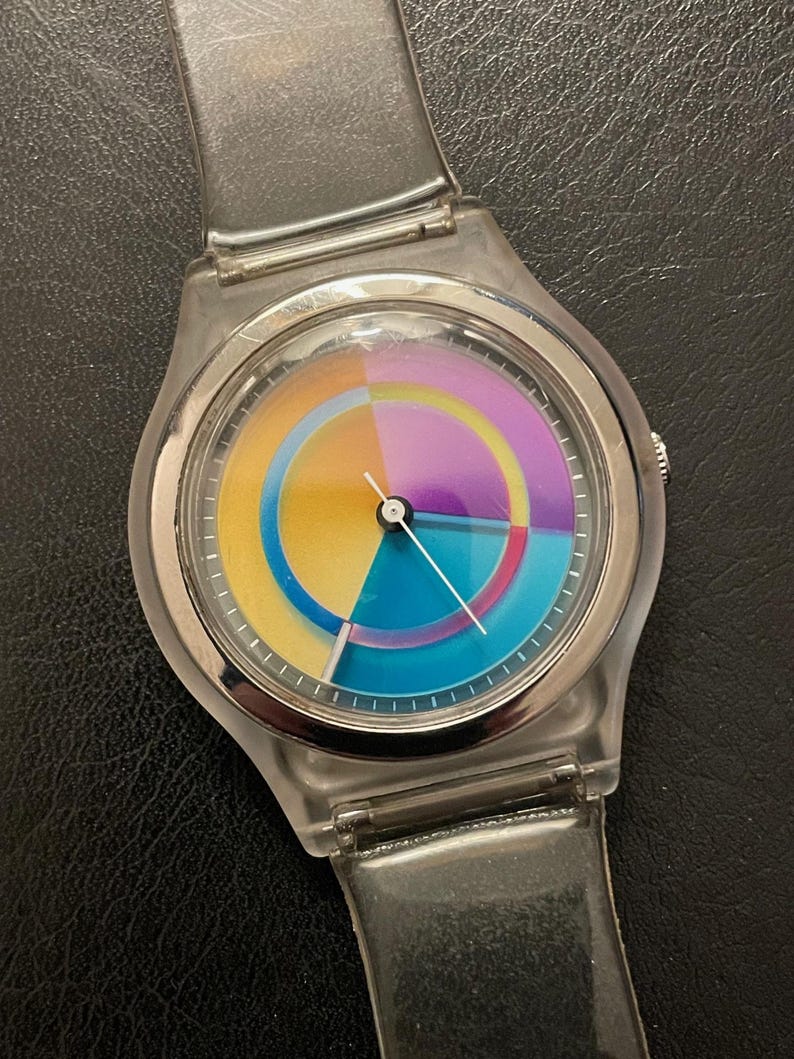May include: A clear plastic wristwatch with a colorful, segmented dial. The dial features a vibrant array of colors including yellow, blue, purple, and orange. The watch has a silver-colored bezel and a clear plastic strap.