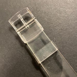 May include: A clear, transparent watch strap with a buckle. The strap is made of a flexible, see-through material. The buckle is silver and has a standard design for securing the watch.