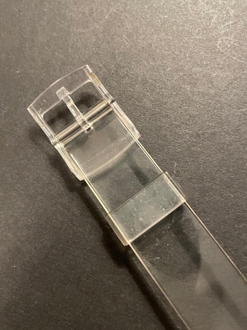 May include: A clear, transparent watch strap with a buckle. The strap is made of a flexible, see-through material. The buckle is silver and has a standard design for securing the watch.