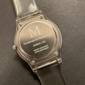 May include: Clear plastic watch with a silver-colored face. The watch face features the text "The Metropolitan Museum of Art" and "MMA.CN". The watch band is clear plastic. The watch is water resistant and has a Japan movement.