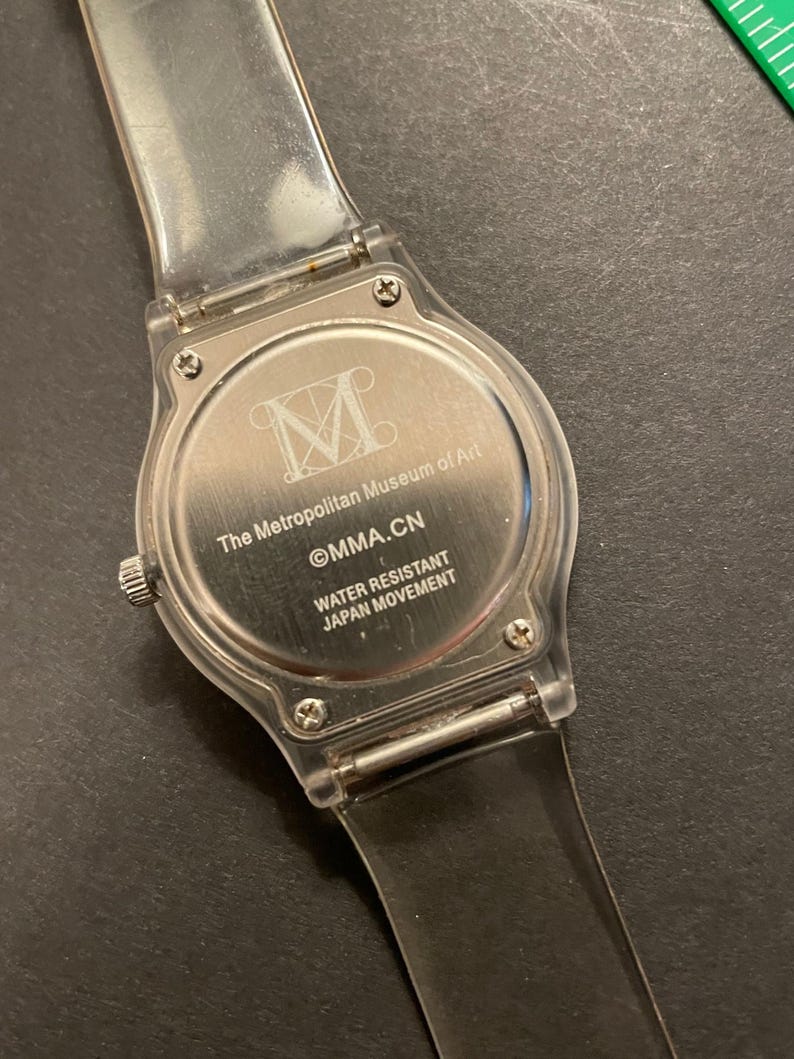 May include: Clear plastic watch with a silver-colored face. The watch face features the text "The Metropolitan Museum of Art" and "MMA.CN". The watch band is clear plastic. The watch is water resistant and has a Japan movement.