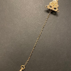 May include: Antique gold-toned watch chain with a decorative triangular top featuring an ornate floral design. The chain is connected to a clasp at the bottom. The watch chain is a vintage accessory.