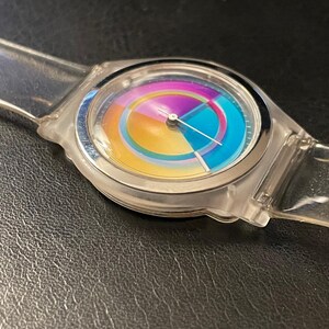 May include: A clear, transparent wristwatch with a colorful rainbow-hued face. The watch has a silver-colored bezel and a clear plastic band. The watch face features a circular design with segments of yellow, orange, blue, and purple. The watch hands are thin and silver.