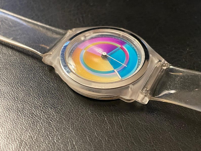 May include: A clear, transparent wristwatch with a colorful rainbow-hued face. The watch has a silver-colored bezel and a clear plastic band. The watch face features a circular design with segments of yellow, orange, blue, and purple. The watch hands are thin and silver.