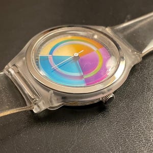 May include: A clear wristwatch with a colorful, segmented face. The watch face features a rainbow of colors, including blue, yellow, purple, and orange. The watch has a clear plastic band and a silver-colored bezel.