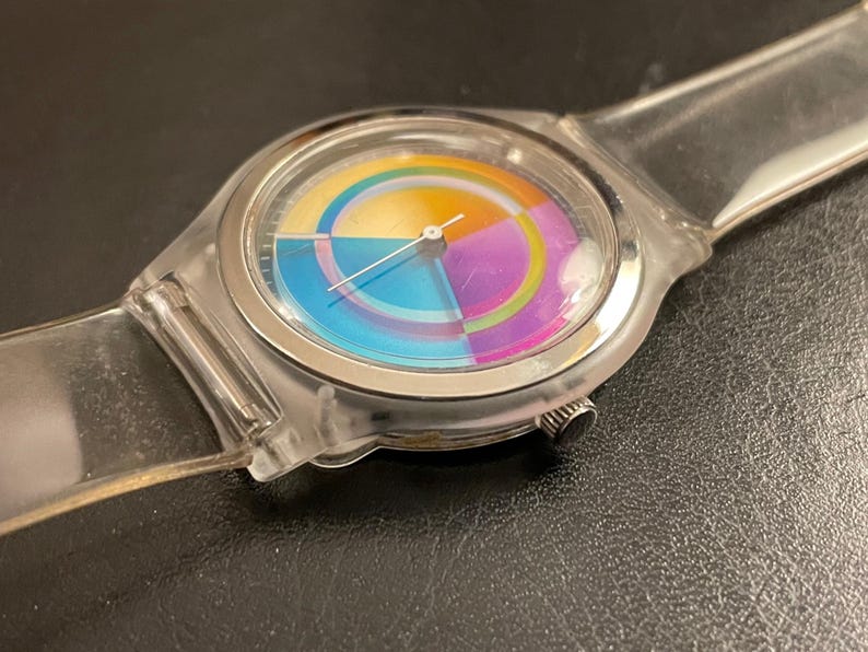 May include: A clear wristwatch with a colorful, segmented face. The watch face features a rainbow of colors, including blue, yellow, purple, and orange. The watch has a clear plastic band and a silver-colored bezel.