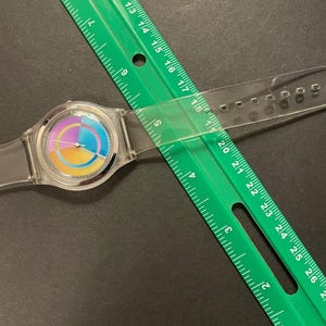 May include: A clear wristwatch with a colorful rainbow-style face is partially covered by a green ruler. The watch has a clear band and a silver-colored bezel. The ruler is marked in inches and is positioned diagonally across the watch.