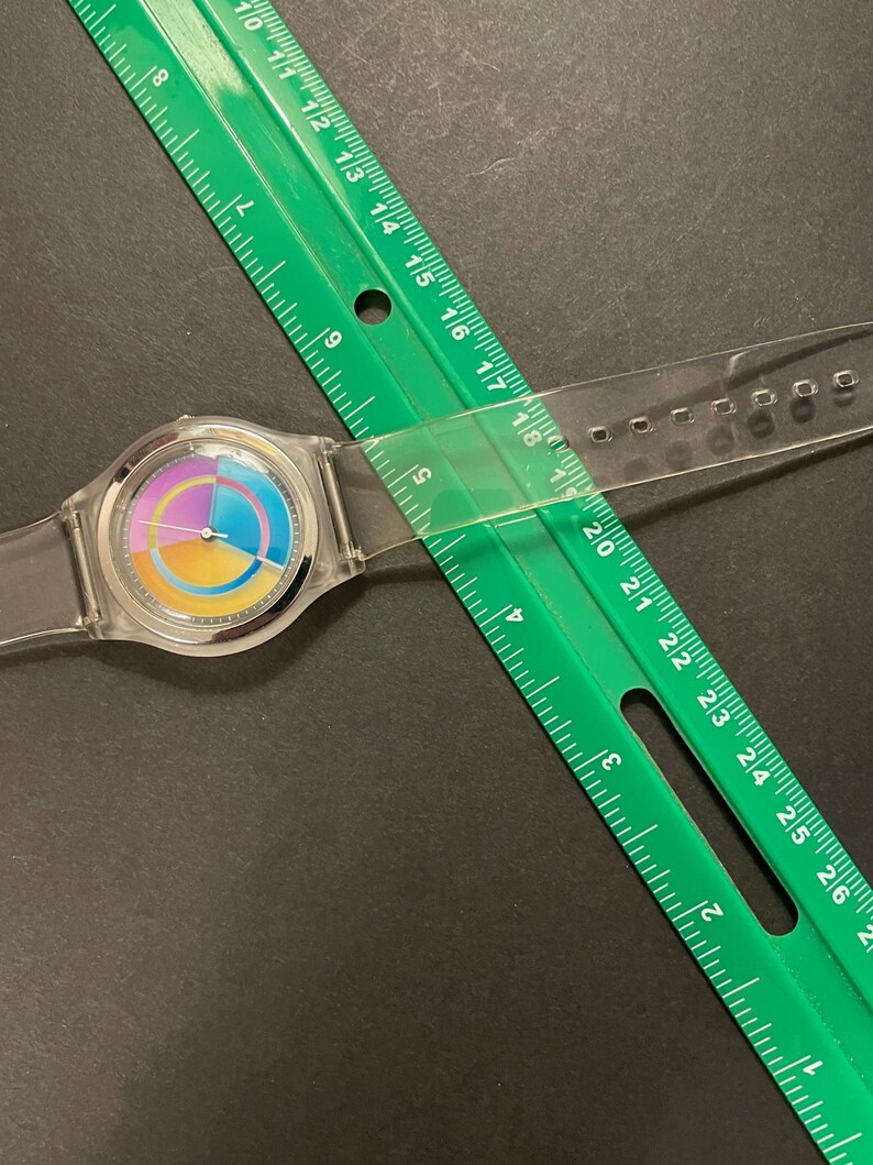 May include: A clear wristwatch with a colorful rainbow-style face is partially covered by a green ruler. The watch has a clear band and a silver-colored bezel. The ruler is marked in inches and is positioned diagonally across the watch.