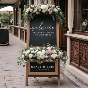 May include: A wooden welcome sign with a black chalkboard surface, adorned with white and pink roses and greenery. The sign displays the word "welcome" and the phrase "WE ARE SO GLAD YOU'RE HERE". The bottom of the sign reads "GRACE & ERIC 7-05-2025".