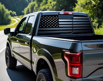 Puede incluir: Una camioneta gris oscuro con un diseño de bandera estadounidense negra en la ventana trasera. La camioneta circula por una carretera con árboles verdes al fondo. La camioneta tiene neumáticos negros y luces traseras rojas.