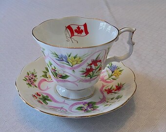 May include: A white teacup and saucer with a floral design and a Canadian flag. The saucer features a pink ribbon with the names of Canadian provinces and territories.
