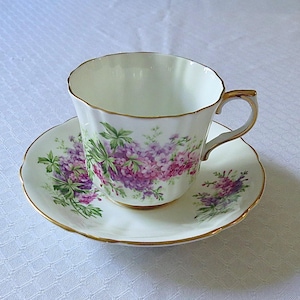 Può includere: Una tazza da tè e un piattino bianchi con un motivo floreale con fiori viola e rosa. La tazza ha un bordo dorato e un manico dorato.