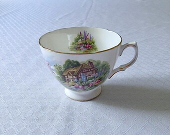 May include: A white teacup with a gold rim and a floral design featuring a cottage with a thatched roof. The design also includes trees, flowers, and a blue sky.