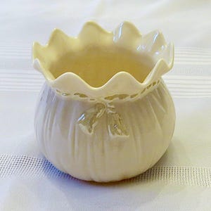 May include: A small, cream-colored ceramic planter with a ruffled rim and a design of a tied bag with a bow. The planter has a textured surface and is perfect for small plants.