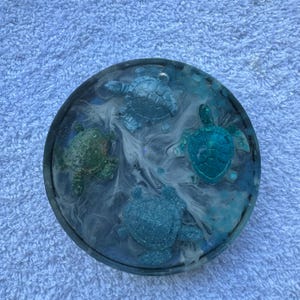 May include: Round resin coaster featuring four sea turtles in shades of blue and green. The turtles are set against a background of swirling white and blue resin, creating an ocean-like effect. The coaster has a dark border.