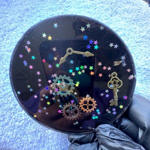 May include: A round, black resin clock with gold clock hands, gears, and a key. The clock face is decorated with colorful star-shaped glitter. The clock is a unique, handmade item.