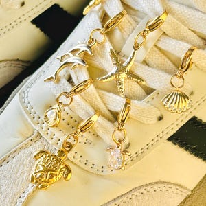 Puede incluir: Abalorios dorados para zapatos con diseños marinos, incluyendo delfines, una estrella de mar, una concha, una tortuga y un pequeño abalorio con una piedra transparente. Los abalorios están sujetos a una zapatilla blanca con detalles crema.