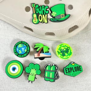 May include: A selection of vibrant shoe charms. The charms include a green guitar with the text "GUAC ON", a green leprechaun hat, a globe with the text "MAKE OUR PLANET GREAT AGAIN", a palm tree, a green portal, a green eye, a broccoli, a scuba tank, and a green mountain with the text "EXPLORE".