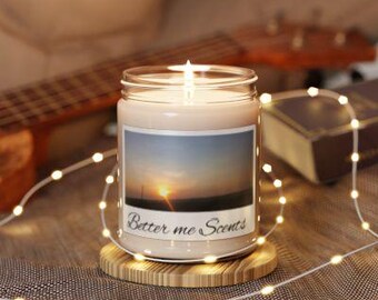 Puede incluir: Un tarro de vela de vidrio transparente con una vela blanca en su interior. La vela tiene una etiqueta que dice "Better Me Scents" y una imagen de una puesta de sol. La vela está sentada sobre una base de madera.