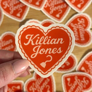 May include: Red heart-shaped stickers with the name "Killian Jones" in white script. The stickers have a scalloped white border and a question mark design. Several stickers are scattered on a wooden surface.