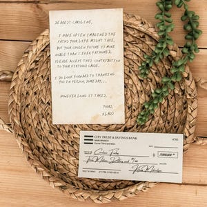 Op de afbeelding: Een handgeschreven brief op verouderd papier met een cheque van drie miljoen euro van City Trust & Savings Bank. De brief luidt: "Lieve Caroline, ik heb me vaak afgevraagd welke paden je leven zou kunnen nemen, maar je gekozen toekomst is nobeler dan ik ooit had kunnen bedenken. Accepteer deze bijdrage voor je edele zaak. Ik kijk ernaar uit om je persoonlijk te bedanken, op een dag... Hoe lang het ook duurt. Met vriendelijke groeten, Klaus."