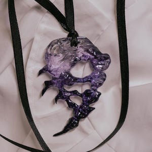 Puede incluir: Un collar con una cinta negra y un colgante de resina transparente y púrpura con forma de diseño abstracto irregular.