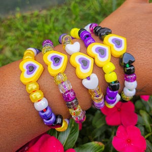 May include: A set of four bracelets with yellow, purple and white beads. Each bracelet features a heart-shaped charm with a yellow, purple and white design. The bracelets are stacked on a person's wrist.