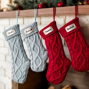 May include: Four cable-knit Christmas stockings in shades of blue and red hang from a wooden mantel. Each stocking has a metallic name tag: "Martha", "Bailen", "Tanner", and "Stella". The stockings are suspended by matching ribbons.