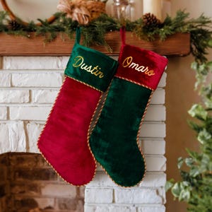 May include: Two Christmas stockings hang on a brick fireplace. One is red with a green cuff and the name "Dustin" embroidered in gold. The other is green with a red cuff and the name "Omaro" embroidered in gold. Both have gold trim.