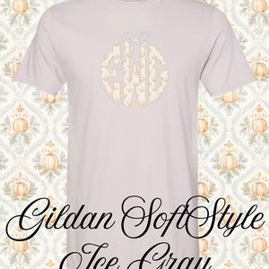 Pode incluir: Uma t-shirt de manga curta cinzento-gelo com um monograma circular com as letras EWG num padrão floral branco e laranja, rematado com um laço branco. O texto Gildan SoftStyle Ice Gray está impresso em letra cursiva preta.