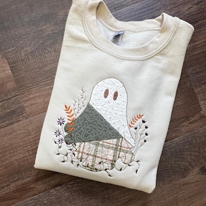 May include: A cream-coloured crewneck sweatshirt featuring an embroidered design of a ghost. The ghost is layered with a dark green quilted fabric and a plaid fabric pattern. Surrounding the ghost are embroidered floral stems in orange and purple.